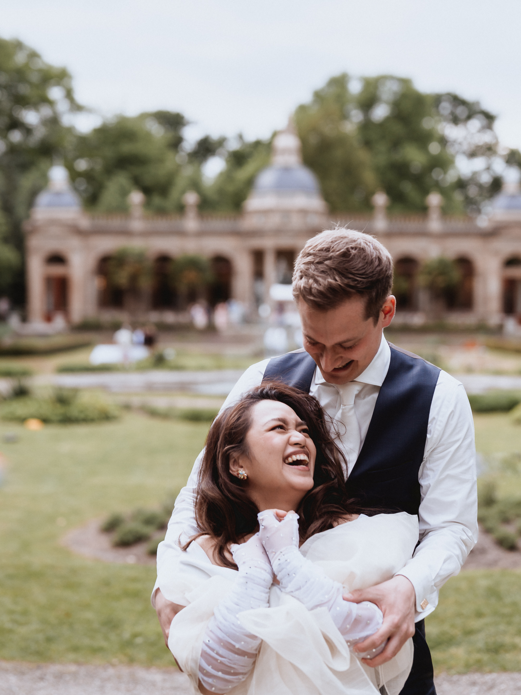 Groom holding his bride from behind while they laugh and smile authentically during their photoshoot at Oranjerie Hydepark in Doorn, captured by Photos by Ville, wedding photographer in the Netherlands for international couples