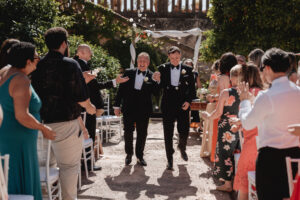 Two grooms walking down the aisle after ceremony with guests celebrating at Finca Son Mir Mallorca photographed by Photos by Ville