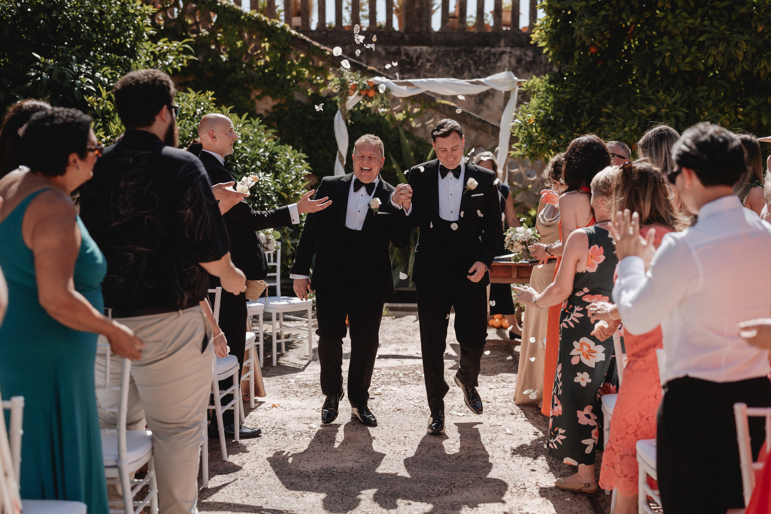 Two grooms walking down the aisle after ceremony with guests celebrating at Finca Son Mir Mallorca photographed by Photos by Ville